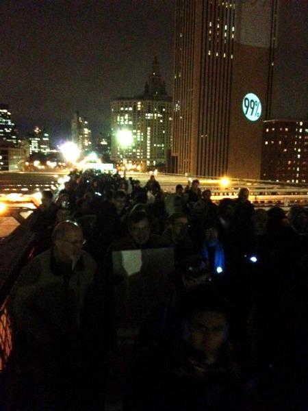 Protesters crossing the Brooklyn Bridge as slogans are projected against nearby buildings.
