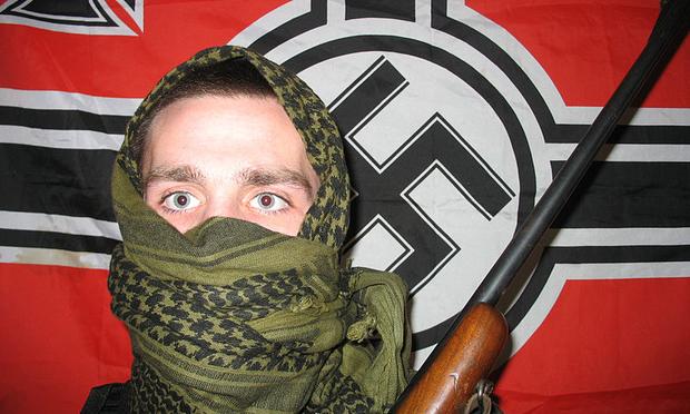 A man holding a rifle in front of a flag with a swastika on it.