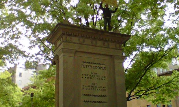 A man scales up the 2-story monument to Peter Cooper carrying a sign that reads 'No Tuition, It's Our Mission.'