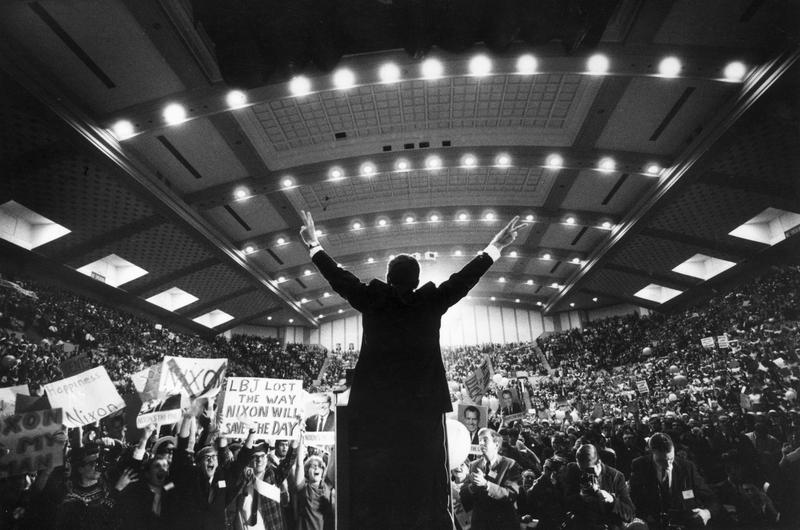 1968: Republican presidential candidate Richard Nixon standing on stage with his back to the camera in front of an arena of supporters giving the victory 'V' sign with both hands.