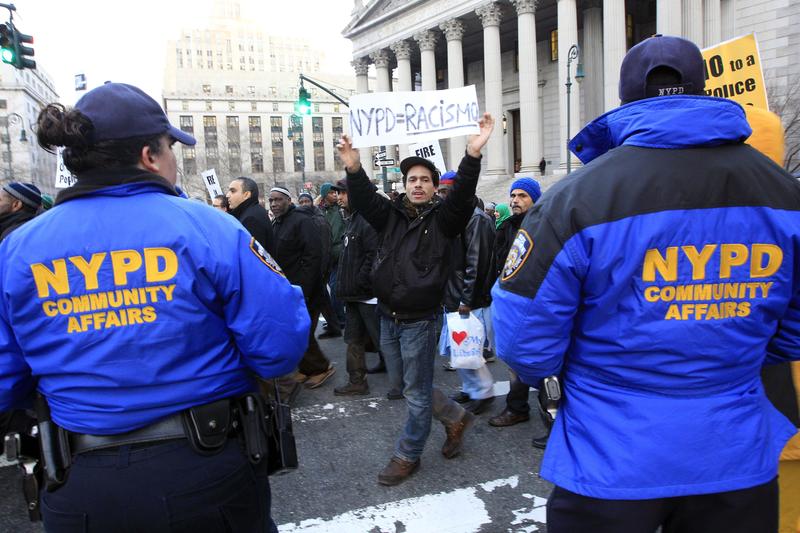 Mariano Munoz, carries a sign past NYPD community affairs officers during rally asking for resignation of NYPD Commissioner R. Kelly & NYPD spokesperson P. Browne, demanding community control of NYPD.