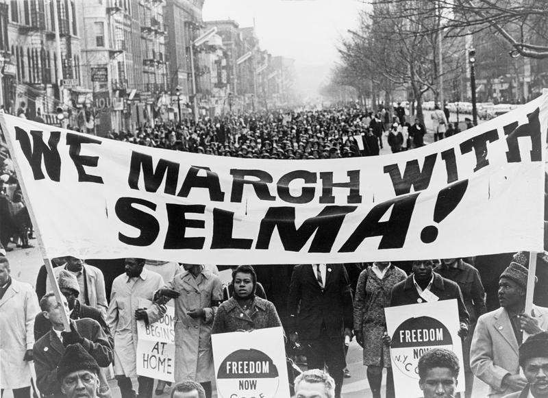 Photograph shows marchers carrying banner "We march with Selma!" on street in Harlem, New York City, New York.
