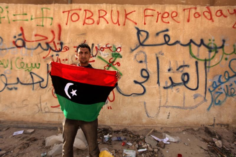 A Libyan anti-government protester holds his old national flag in front of a wall covered with graffiti against Libyan leader Moamer Kadhafi in the eastern city of Tobruk on February 24, 2011.