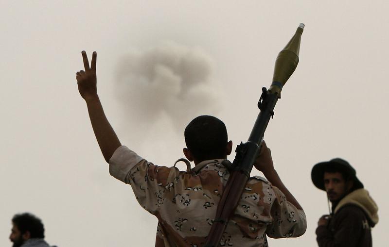 A Libyan rebel walking with a rocket-propelled grenade (RPG) flashes the victory sign as smoke billows from a distance in the eastern city of Ajdabiya on March 30, 2011.